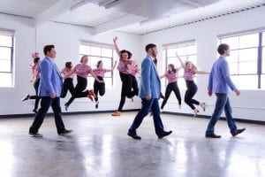Picture of dancers jumping in warehouse, with men walking in front 