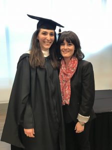 Woman in graduation regalia embraces woman in black blazer and pink scarf.