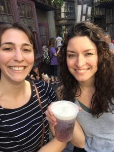 Two women with brown hair pose for a picture at Harry Potter World. One holds a cup of butter beer.