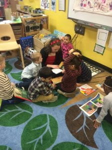 Teacher sitting on carpet reading to 5 Children around her in a circle. 