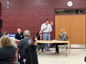 Four adults sit at a table and hold microphones and a script in their hands. They are performing a skit about mental health.