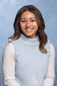Portrait-style photo of Alex Coyne smiling at the camera, wearing a white shirt and blue sweater in front of a light blue background