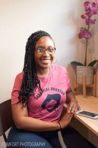 Sharbreon sits at a desk, turned toward camera, with pink Zora Neal Hurston t-shit