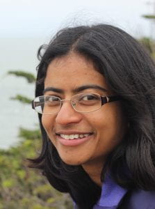 Dr Amrita Dhar, a South Asian woman wearing a purple collared shirt and eyeglasses. She stands on a beach with greenery lining the shore.