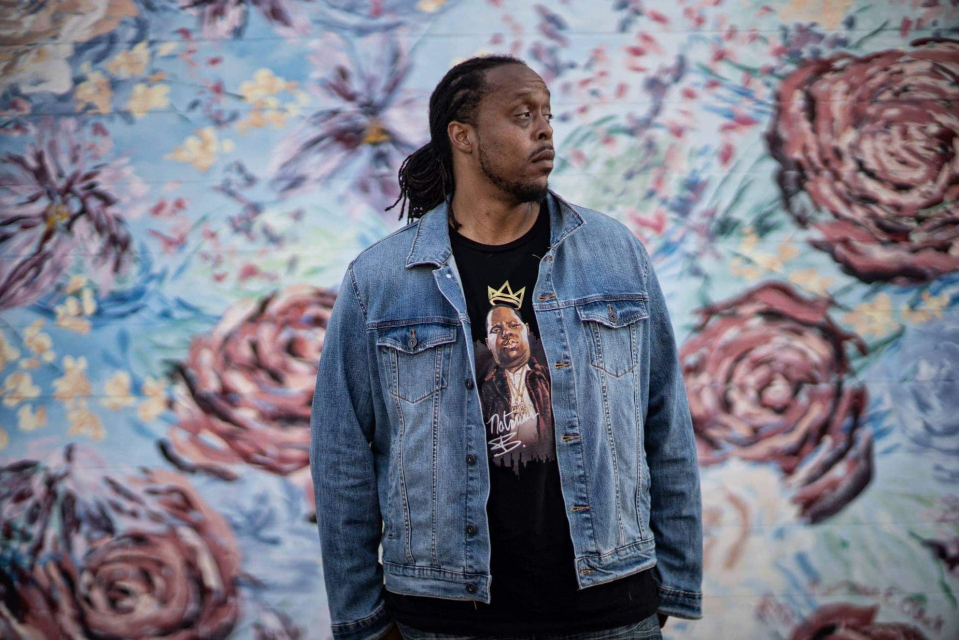 Evan Williams, an African-American man, wearing a blue denim jacket and black shirt featuring a portrait of The Notorious B.I.G. He is wearing his dreadlocks tied back and is looking to his left. Behind him is a colourful mural of various kinds of flowers.