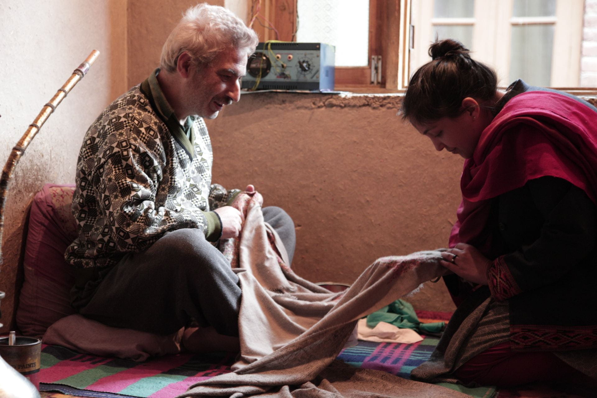 A Kashmiri man and woman sit next to each other on the ground while working on sewing a piece of fabric. 