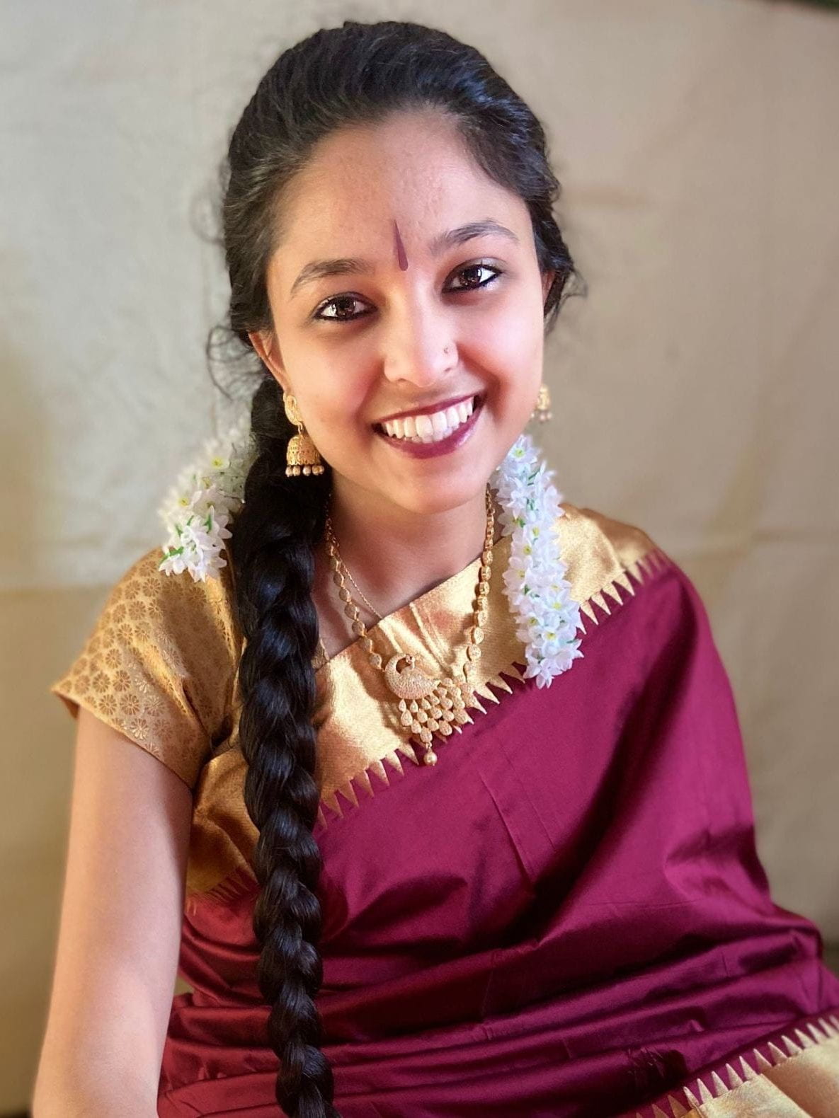 Sushmitha Ravikumar, a South Asian woman in a gold-bordered maroon sari and wearing her long dark hair braided over her shoulder, smiles as she sits in front of a taupe-coloured wall.