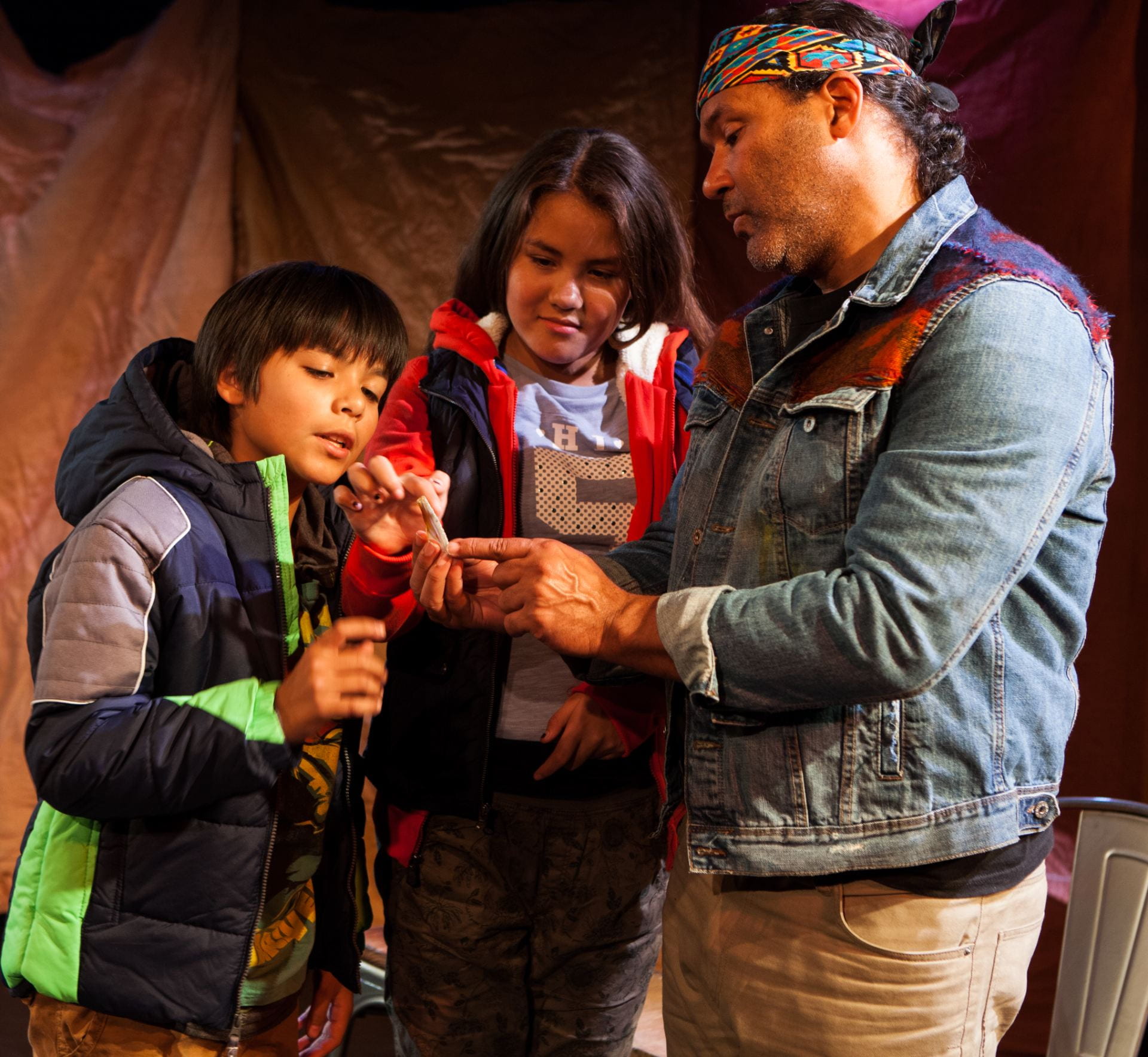 Production still from Amerinda, INC.'s adaptation of Powwow Highway, adapted by William S. Yellowrobe, Jr. and directed by Madeline Sayet. Image depicts an an older man talking with two young kids as they examine an object in the man's hand. 