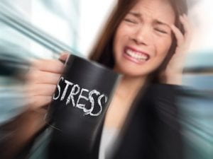 A picture of a lady holding one hand to her head while grimacing, as the other hand is holding a coffee cup that says “stress” on it