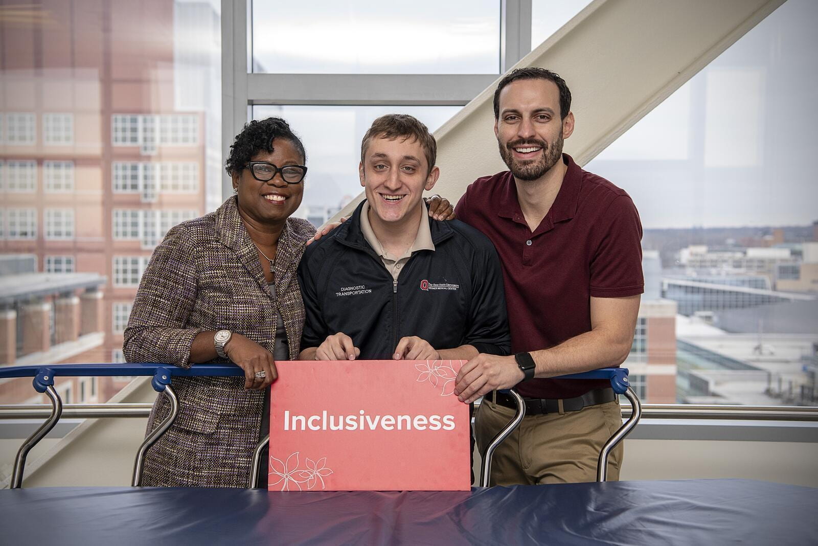 Ben and his two supervisors smiling in front of a patient transportation cart. They are holding a red sign that says "Inclusiveness"