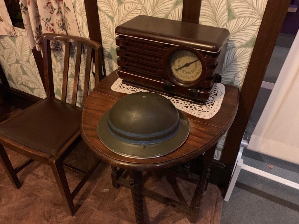 A World War II era helmet sits atop a table. The radio next to it plays a BBC broadcast declaring Labour's landslide victory in 1945.