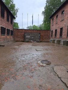A largely empty, concrete and brick courtyard between two buildings. There is a wall at the far end with flowers in front of it as a memorial to the prisoners the Nazis executed there.