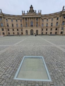A photo of the Empty Library memorial. The memorial is a room filled with empty bookshelves in the ground, covered by a sheet of glass. In the background is Humboldt-Universität.